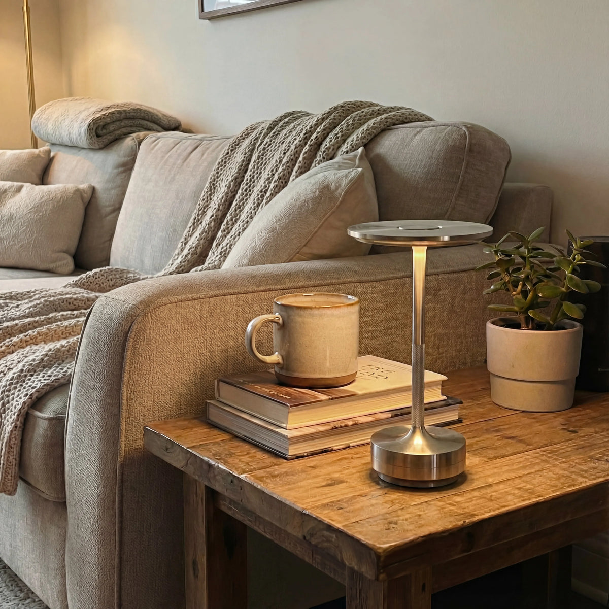 Gold Eclara Ilumina rechargeable table lamp on a wooden table in a living room with beige sofa, ochre mug and books